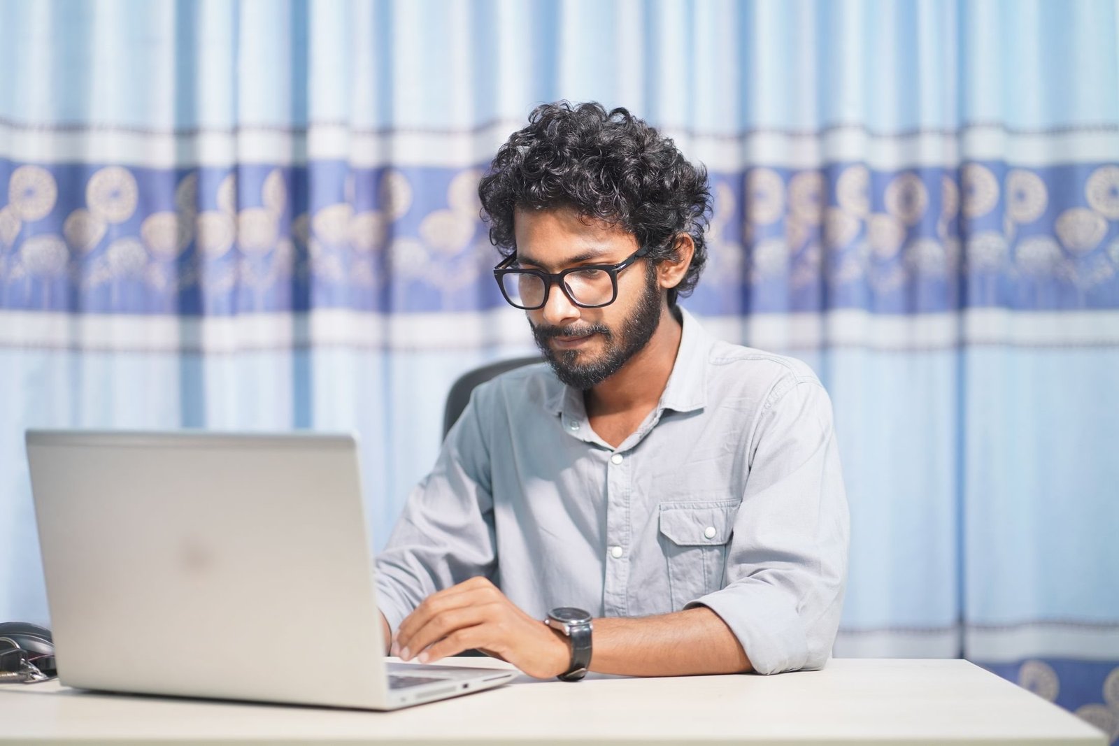 A developer with glasses and a beard works on a laptop at a desk in front of a patterned curtain