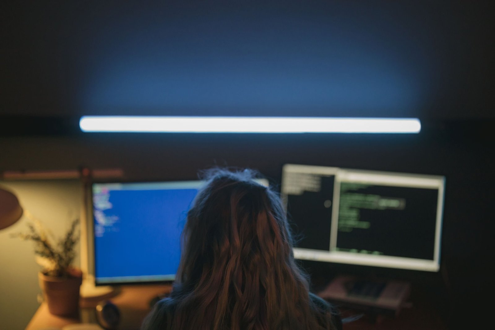 A developer seen from behind works at two monitors in a dim room lit by a strip light