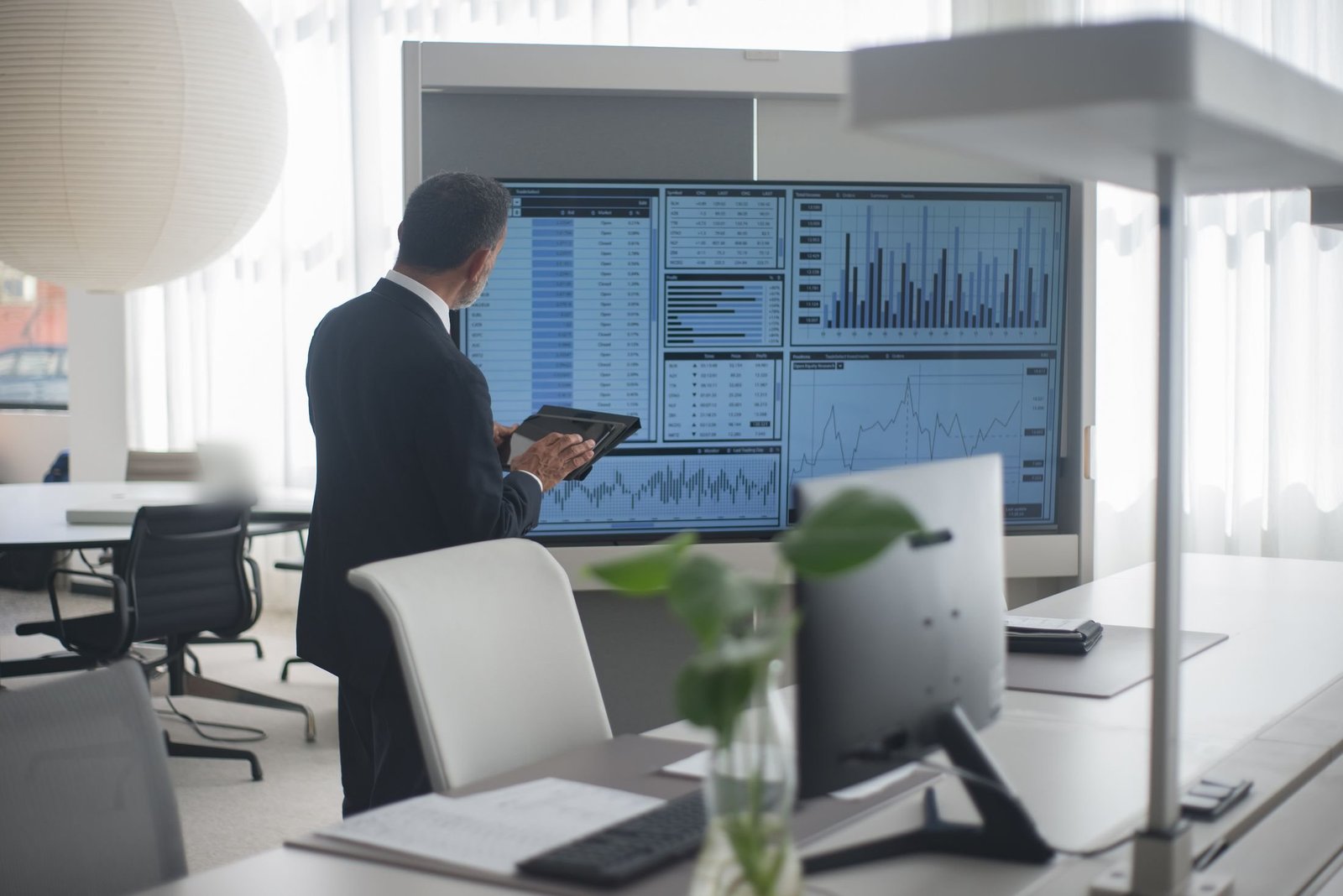 Someone in a suit studies a wall-sized dashboard of charts and tables while holding a tablet in a bright office