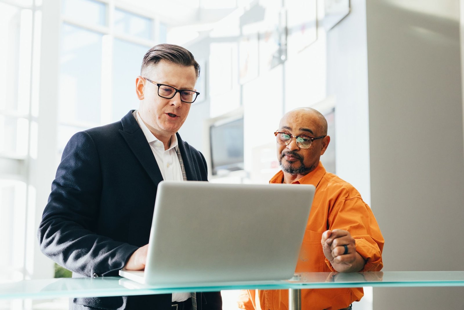 Two colleagues stand at a glass table discussing something on a laptop screen