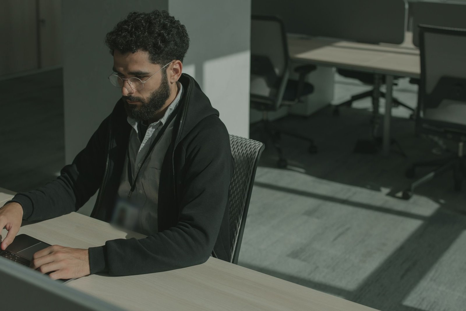 A developer in a dark hoodie with a lanyard types on a laptop at a desk in a quiet office