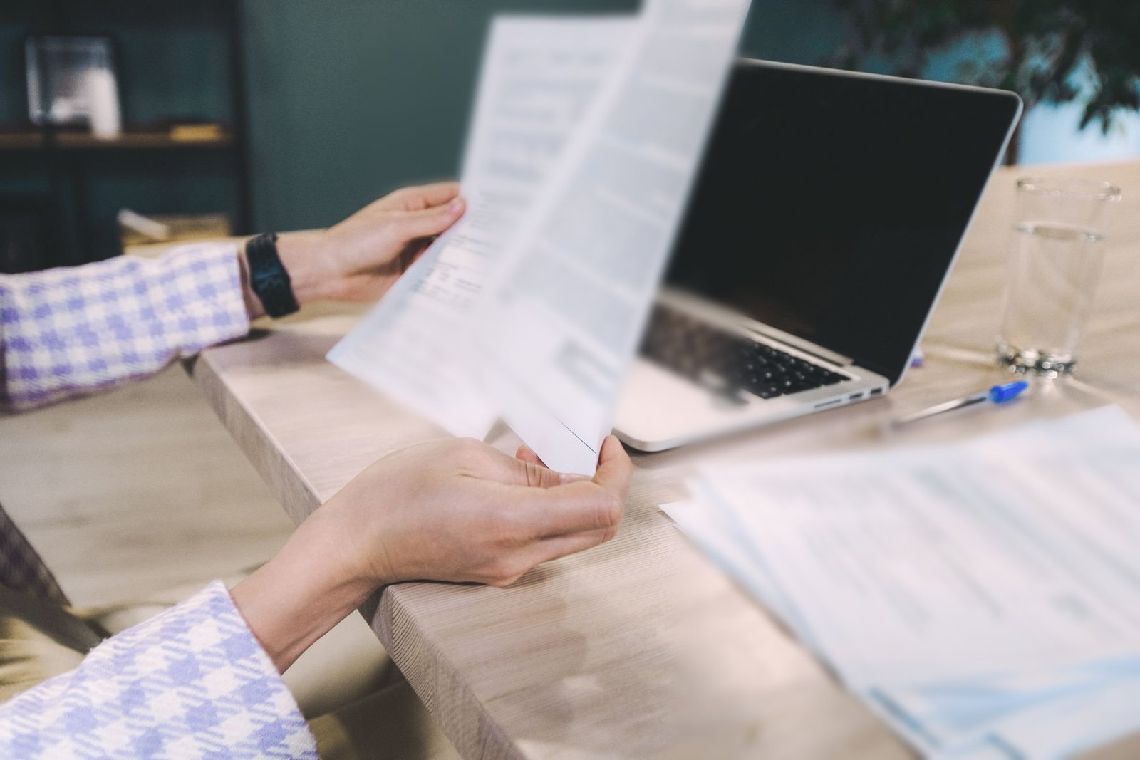 Hands hold two printed forms beside an open laptop, more paperwork stacked on the desk